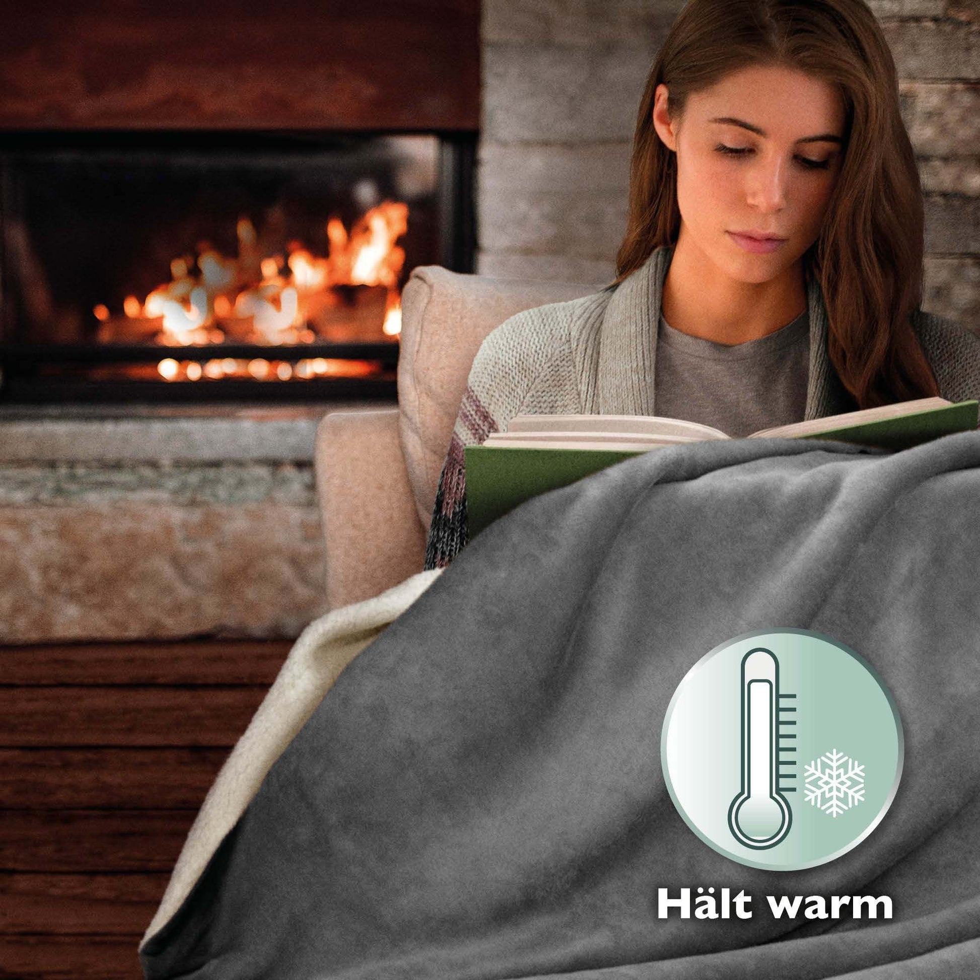 Cozy woman wrapped in Beautissu Sherpa Kuscheldecke Alina blanket by fireplace, reading.