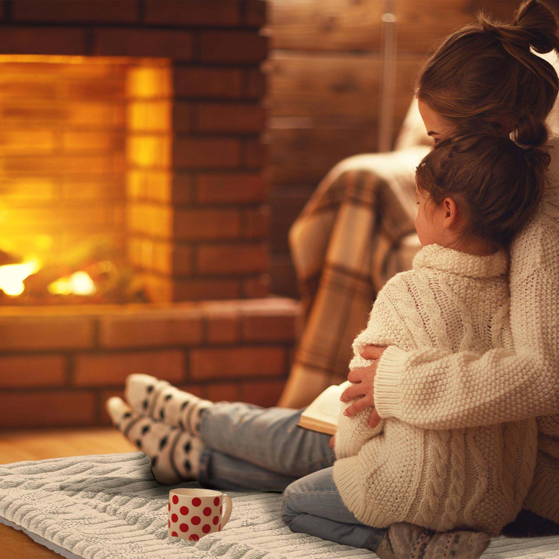 Cozy scene with a mother and child in sweaters sitting on a fluffy blanket by a fireplace, enjoying warmth and comfort.